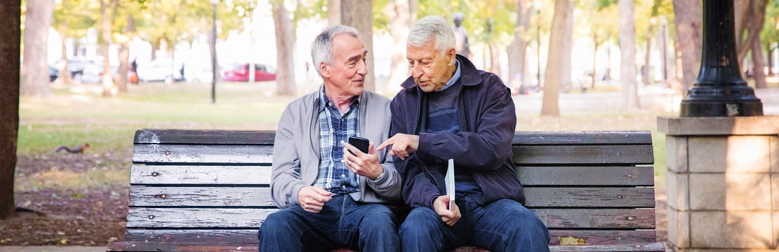 An elderly same sex couple sits on a park bench talking.